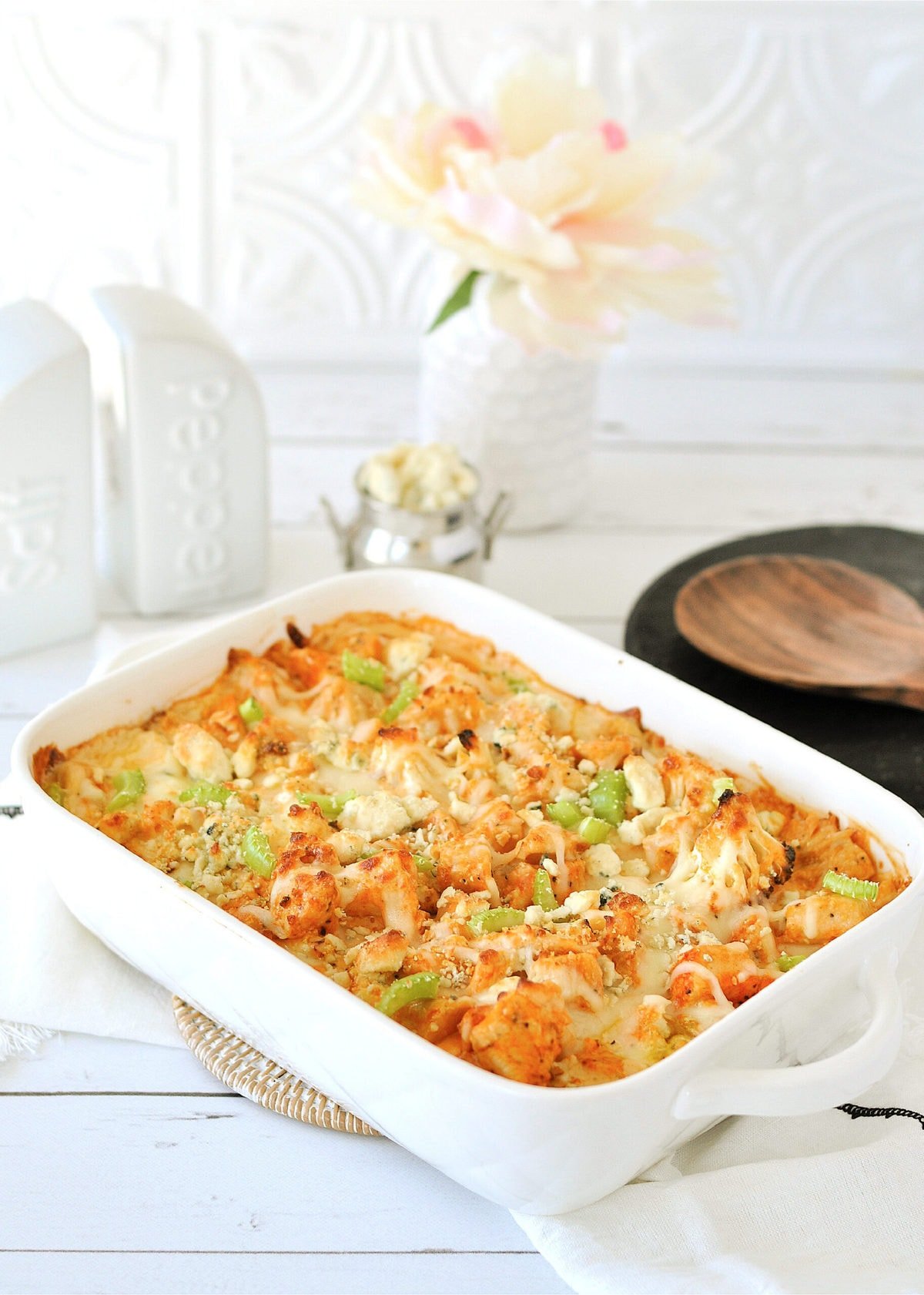 a white casserole dish, filled with buffalo chicken cauliflower Mac and cheese, with a white cloth and wooden serving spoon