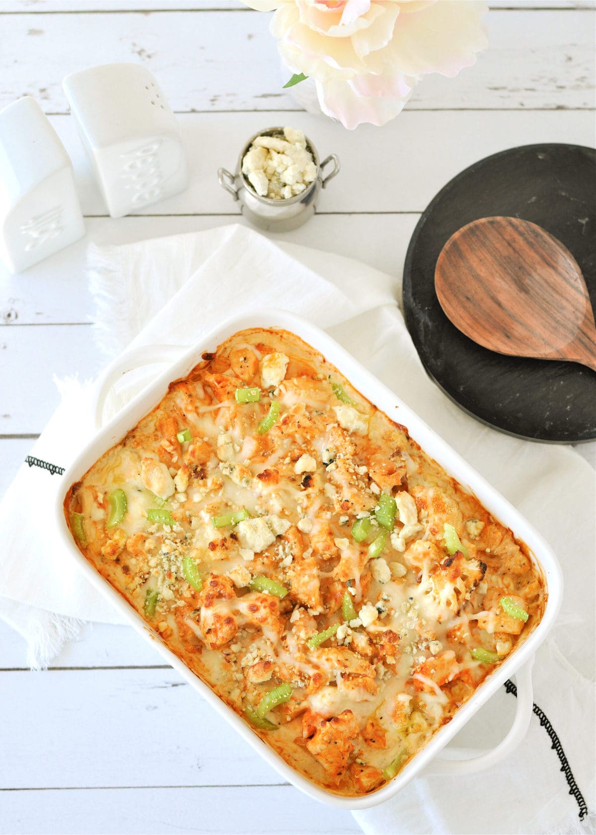 a white casserole dish, filled with buffalo chicken cauliflower Mac and cheese, with a white cloth and wooden serving spoon
