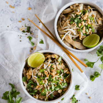 Two bowls of Keto Chicken Pad Thai garnished with red pepper flakes, cilantro, and lime in white bowls on a white background with wooden chopsticks and extra garnish sprinkled around.