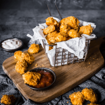 Keto Popcorn Chicken in a frying basket lined with a white napkin, on a wood cutting board with barbecue sauce and ranch to the side.