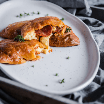 Keto pepperoni pizza pockets, sliced in half piled on top of each other with gooey cheese and pepperoni oozing out on top of a white plate. Fresh herbs are sprinkled around the plate, which sits on a light gray fabric napkin.