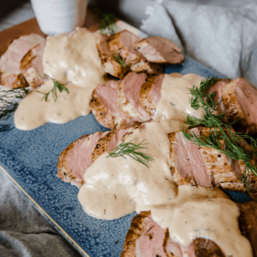 Pork Tenderloin sliced on a blue ceramic cutting board and topped with creamy dill sauce, garnished with fresh dill.