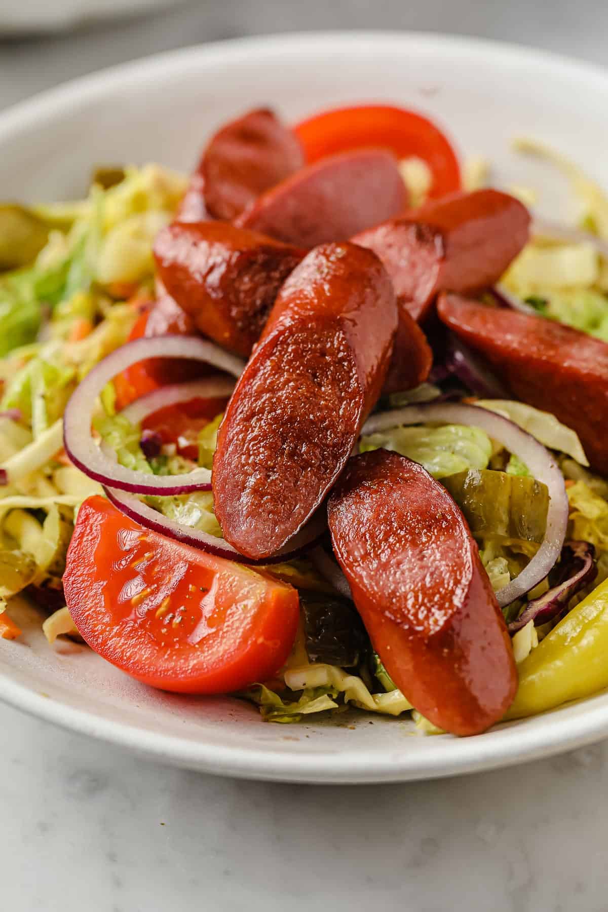 a salad piled high with grilled hot dogs, pickles, red onion, peppers, tomatoes, and a mustard dressing