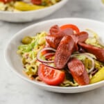 a salad piled high with grilled hot dogs, pickles, red onion, peppers, tomatoes, and a mustard dressing.