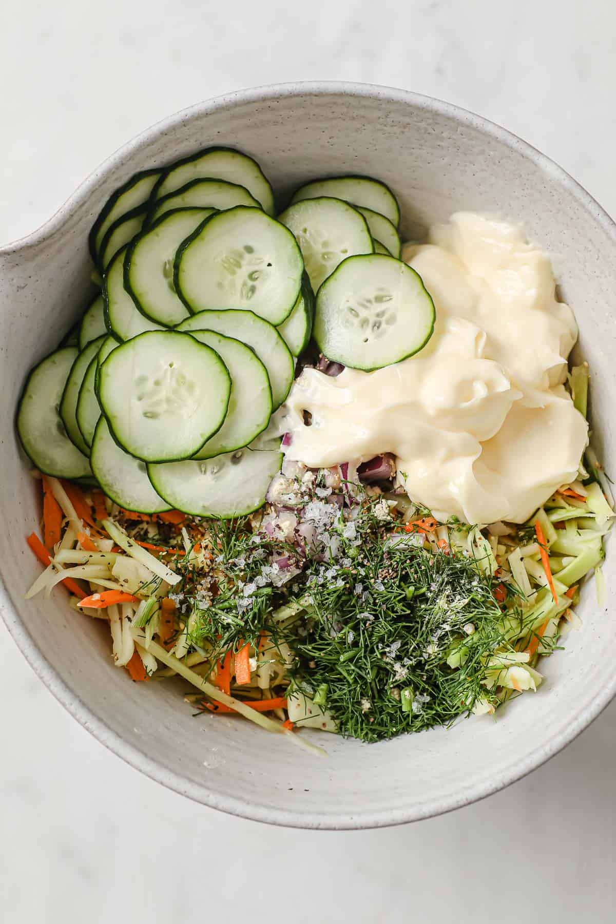 Ingredients for keto coleslaw in a mixing bowl - broccoli slaw, cucumbers, mayo, vinegar, dill, garlic, salt and pepper