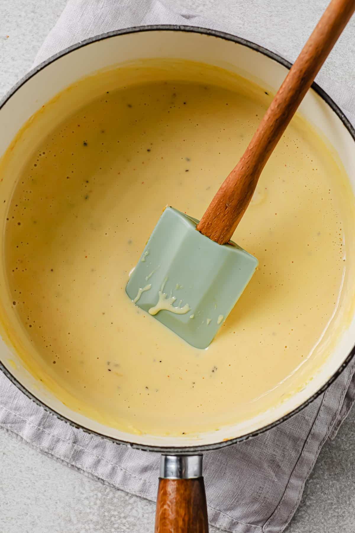 a white sauce pan with a melty cheese sauce inside, with a rubber spatula