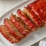meatloaf topped with ketchup, sliced and served on a white serving platter - gravy and mashed cauliflower in the background