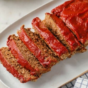 meatloaf topped with ketchup, sliced and served on a white serving platter - gravy and mashed cauliflower in the background