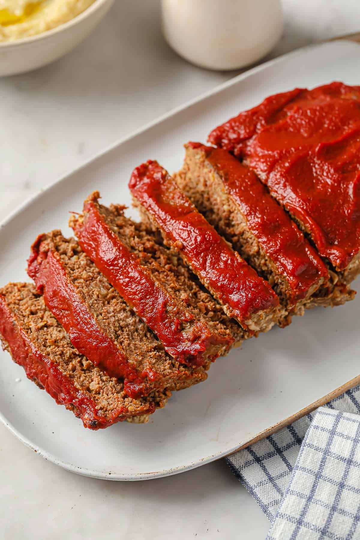 meatloaf topped with ketchup, sliced and served on a white serving platter - gravy and mashed cauliflower in the background