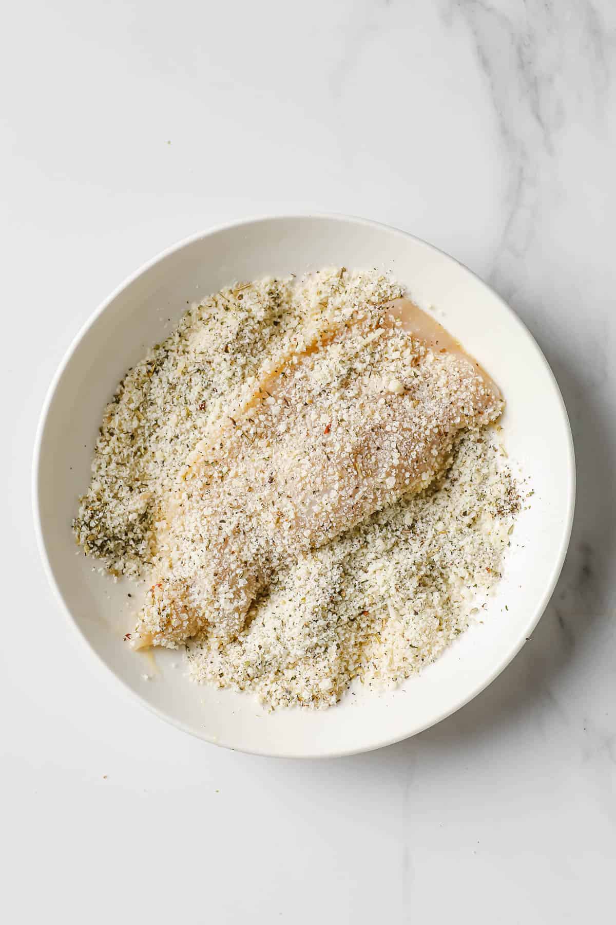 a white bowl with a raw chicken breast being coated in parmesan cheese and herbs