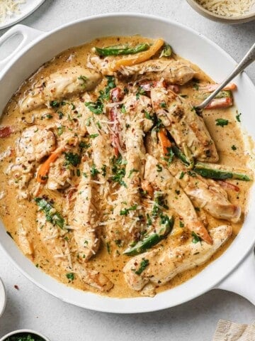 a white skillet with sautéed chicken, creamy sauce, peppers and onions, garnished with cheese and parsley