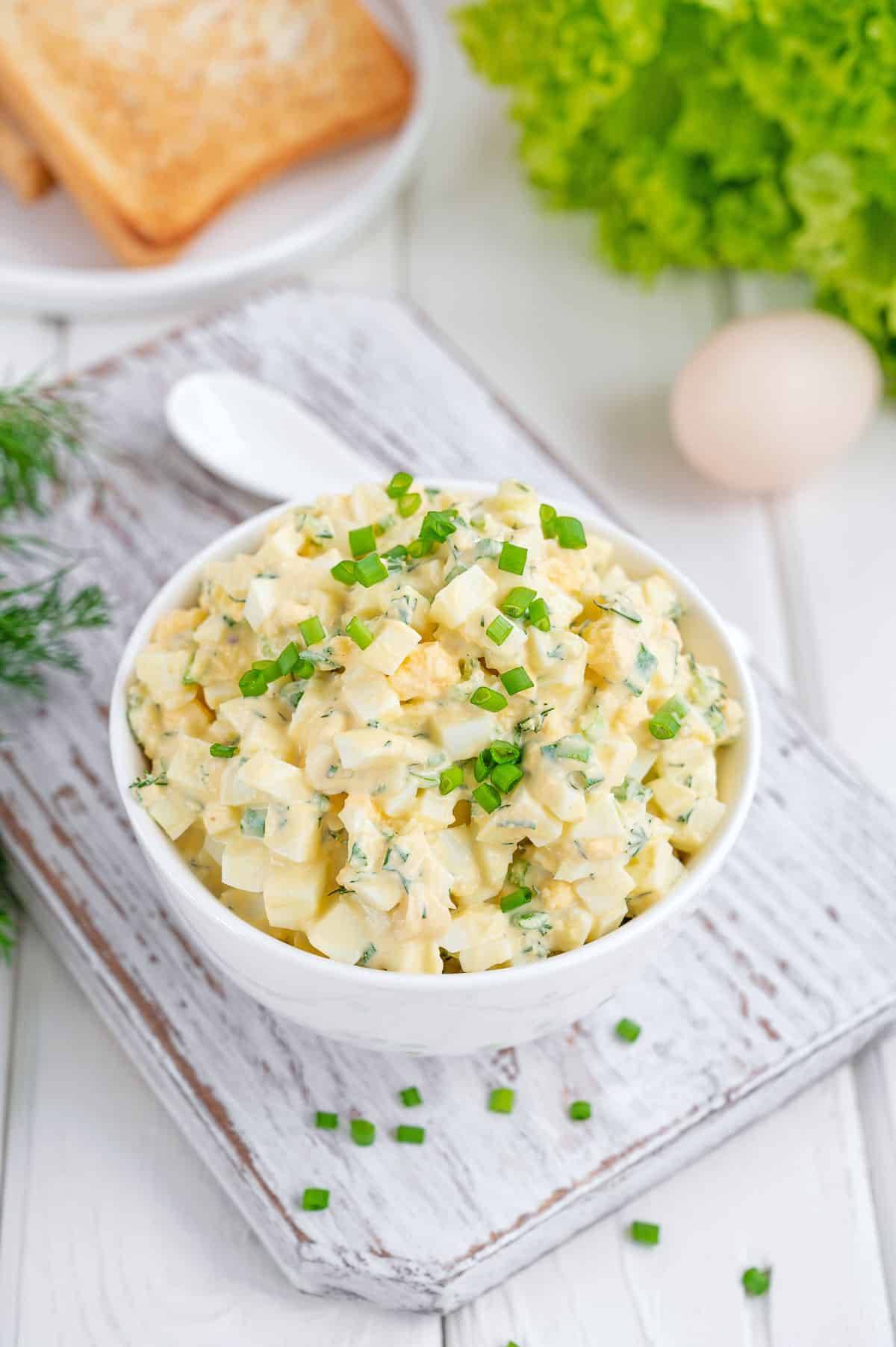 a white bowl full of egg salad, garnished with green onions, with tomato, bread and eggs in the background