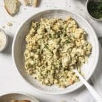 A bowl of egg salad, surrounded by roasted garlic, parsley and crostini.