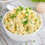 a white bowl full of egg salad, garnished with green onions, with tomato, bread and eggs in the background