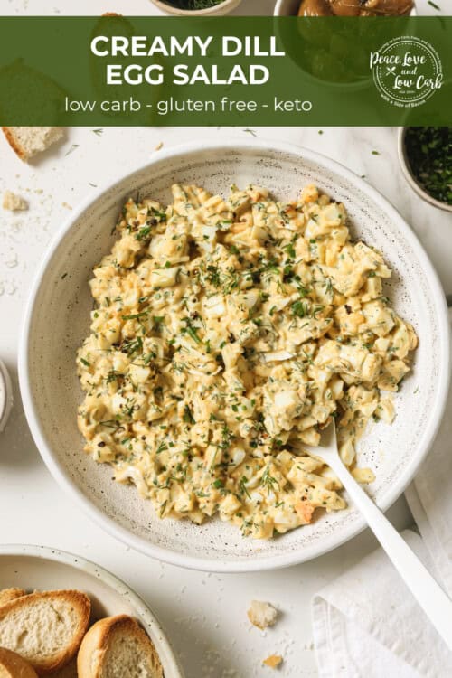 A bowl of egg salad, surrounded by roasted garlic, parsley and crostini.