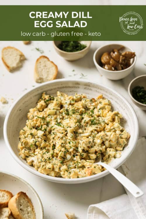 A bowl of egg salad, surrounded by roasted garlic, parsley and crostini.