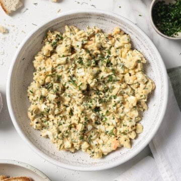 A bowl of egg salad, surrounded by roasted garlic, parsley and crostini.