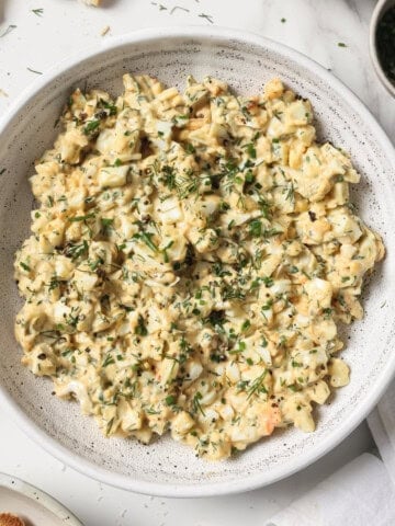A bowl of egg salad, surrounded by roasted garlic, parsley and crostini.