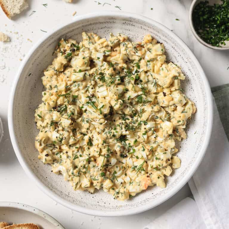 A bowl of egg salad, surrounded by roasted garlic, parsley and crostini.