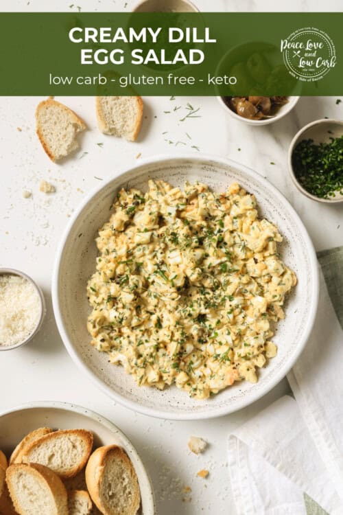 A bowl of egg salad, surrounded by roasted garlic, parsley and crostini.