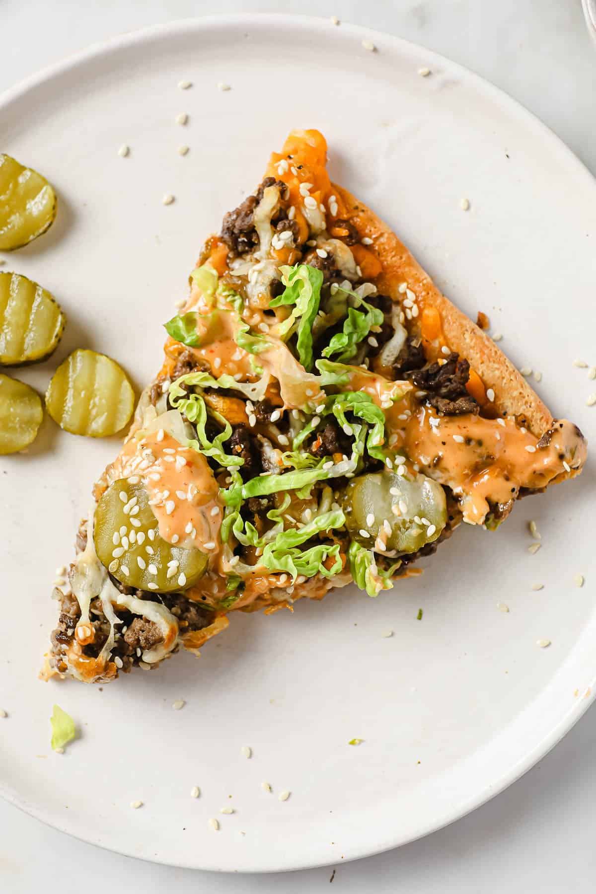a slice of low carb Big Mac pizza on a white plate, with a side of pickles.
