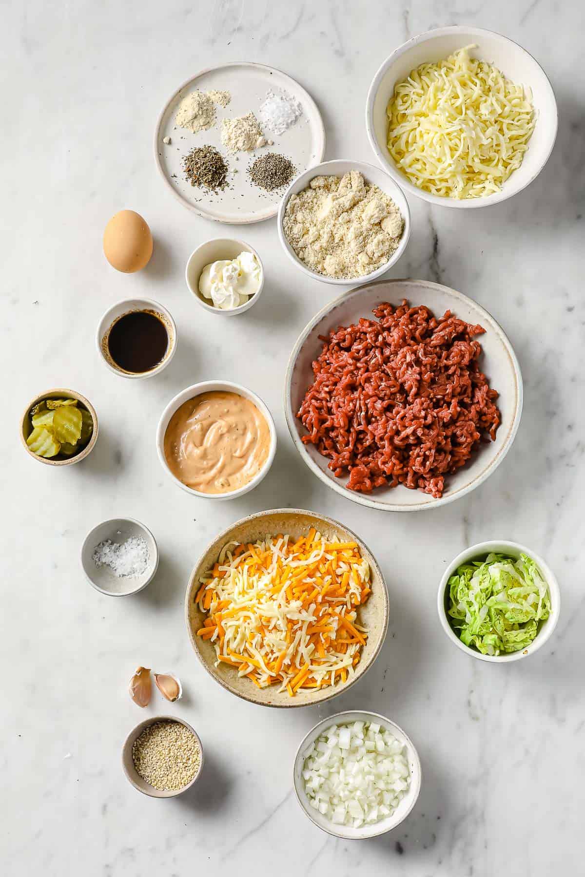 Ingredients laid out in individual bowl - ground beef, cheese, lettuce, sauce, onions, pickles, sesame seeds.