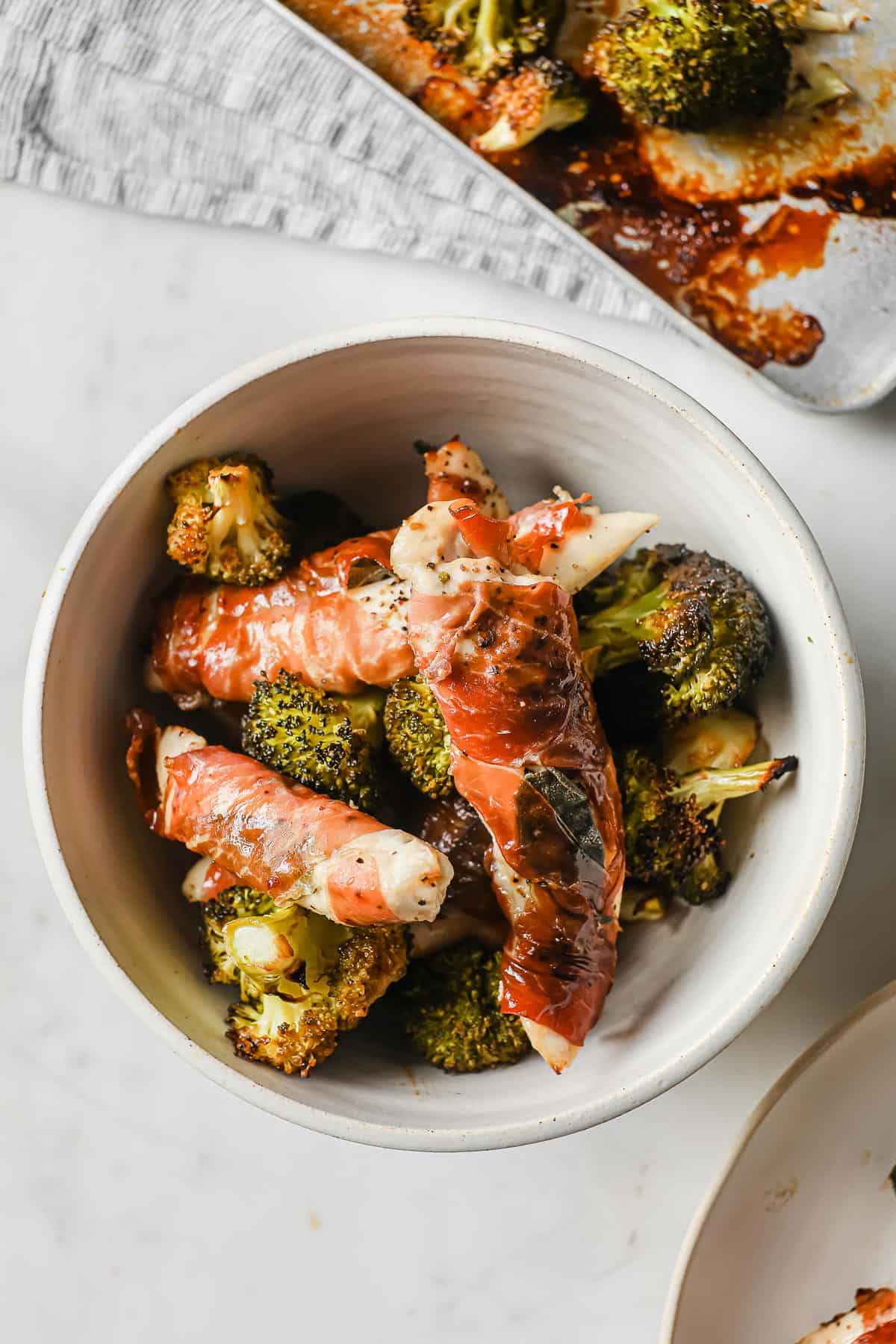 a white bowl with roasted broccoli and prosciutto wrapped chicken.