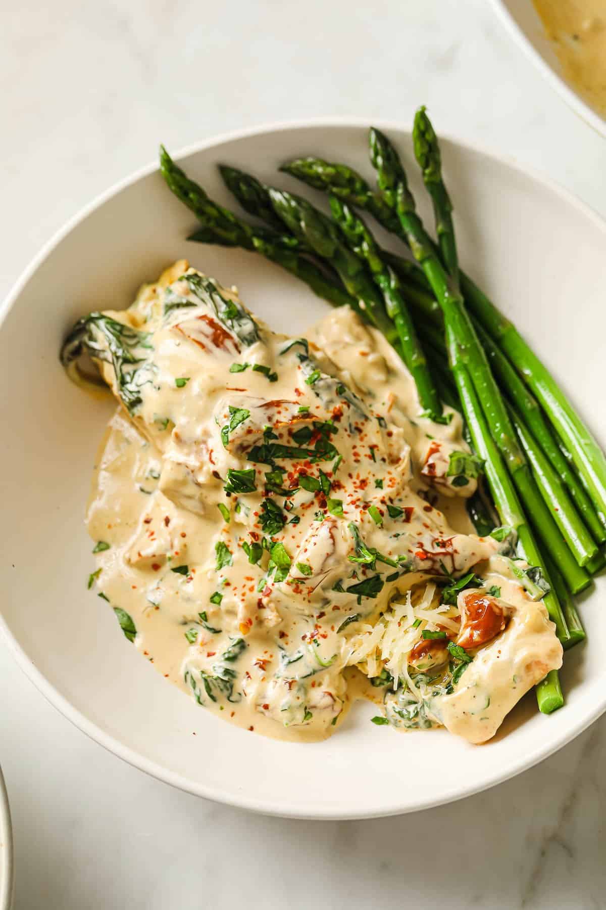 a white bowl with a portion of creamy tuscan salmon, served with steamed asparagus.