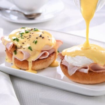 a white plate with a serving of eggs Benedict, with a gravy boat and a basket of buns in the background.
