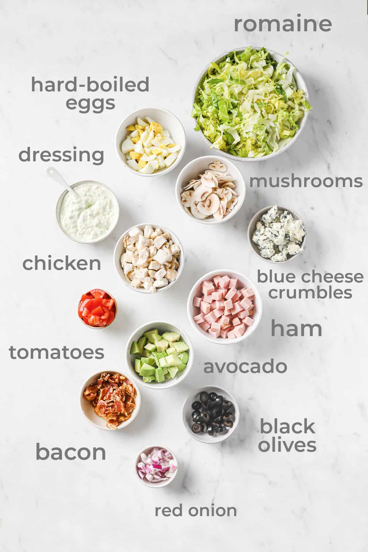 Ingredient laid out to make a keto Cobb salad - lettuce, ham, chicken, bacon, tomatoes, mushrooms, red onion, cheese, and dressing.