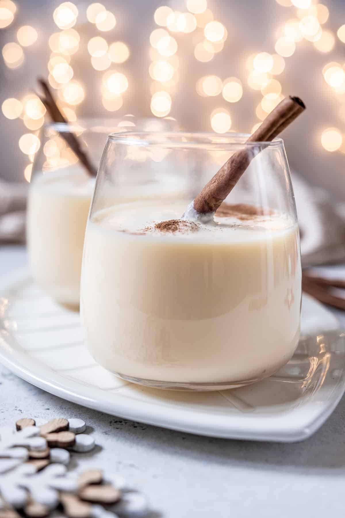 Two stemless wine glasses full of homemade eggnog, garnished with nutmeg and a cinnamon stick.