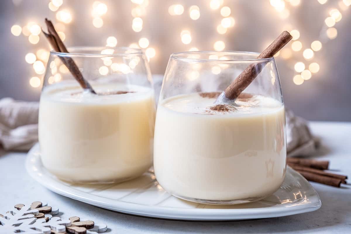 Two stemless wine glasses full of homemade eggnog, garnished with nutmeg and a cinnamon stick.