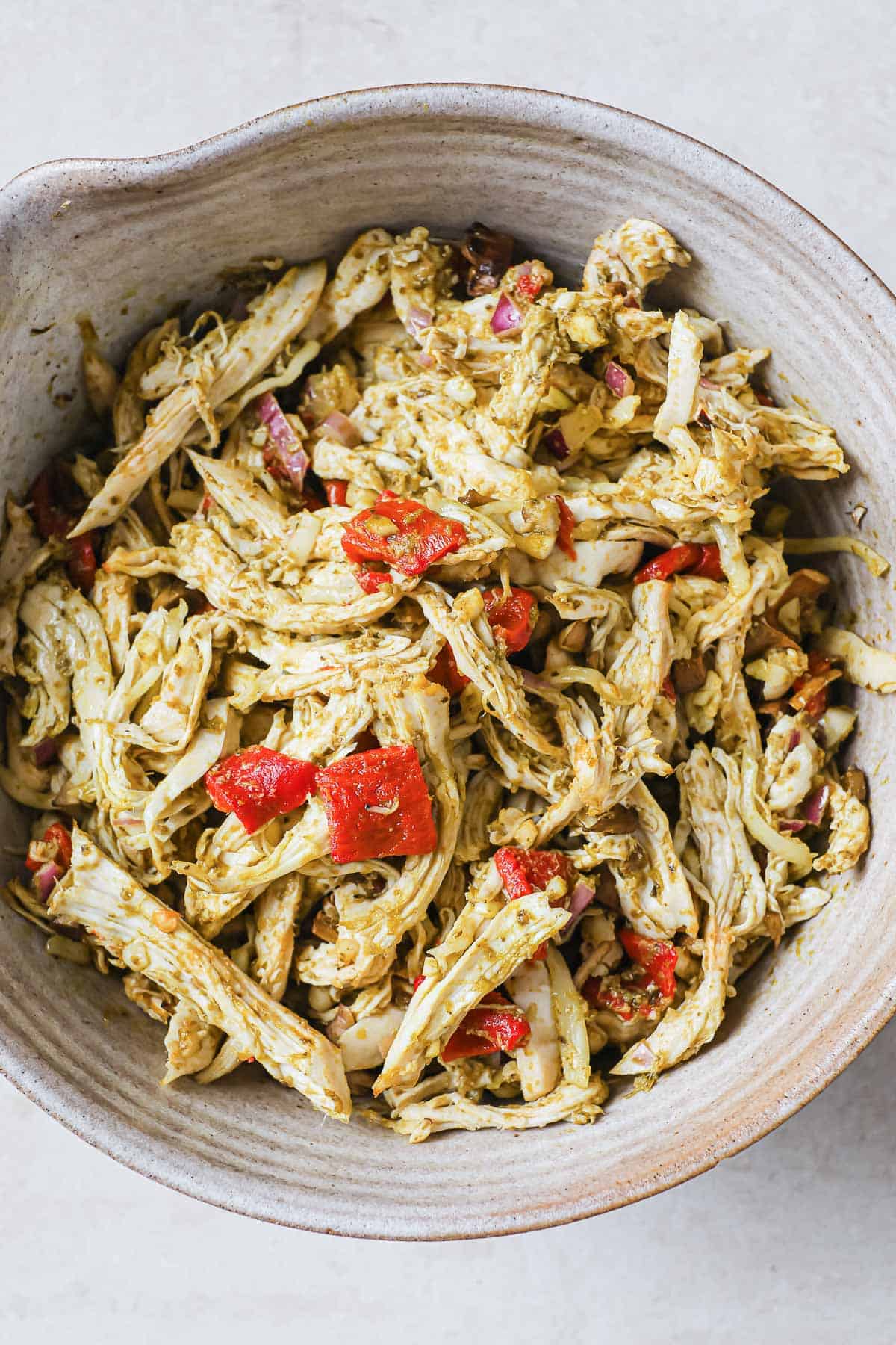 A mixing bowl filled with shredded chicken, tossed in pesto, with roasted red peppers and red onions.