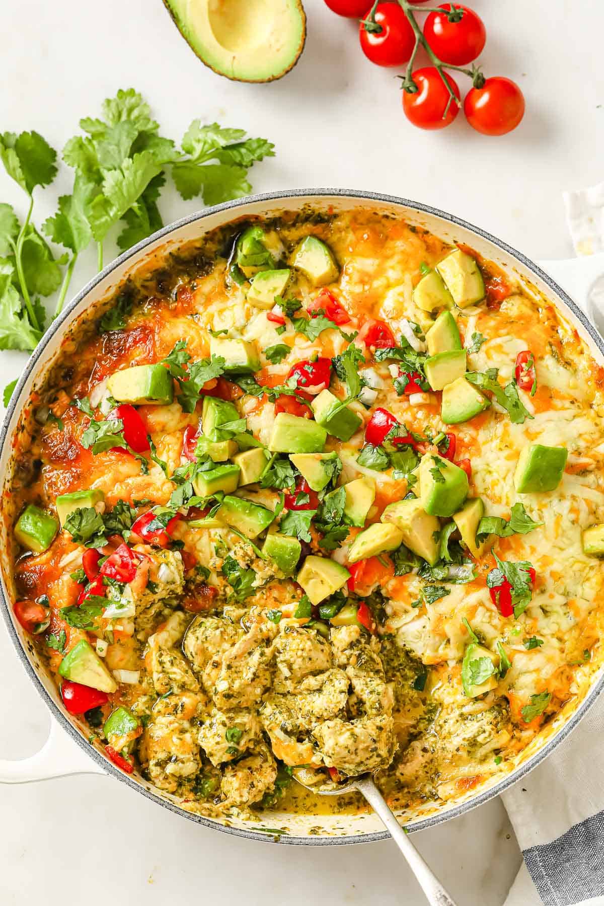 An enameled cast iron skillet filled with a cheesy chicken casserole, topped with salsa verde, cheese, pico de Gallo, avocado, and cilantro.