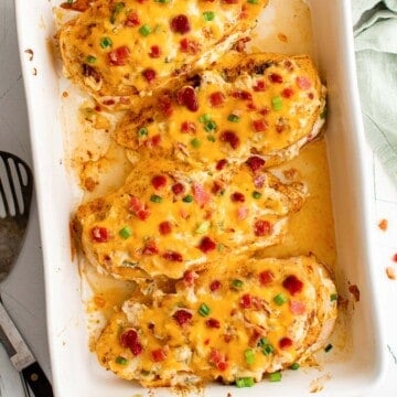 A white casserole dish with chicken breasts topped with cheese, bacon and green onions.