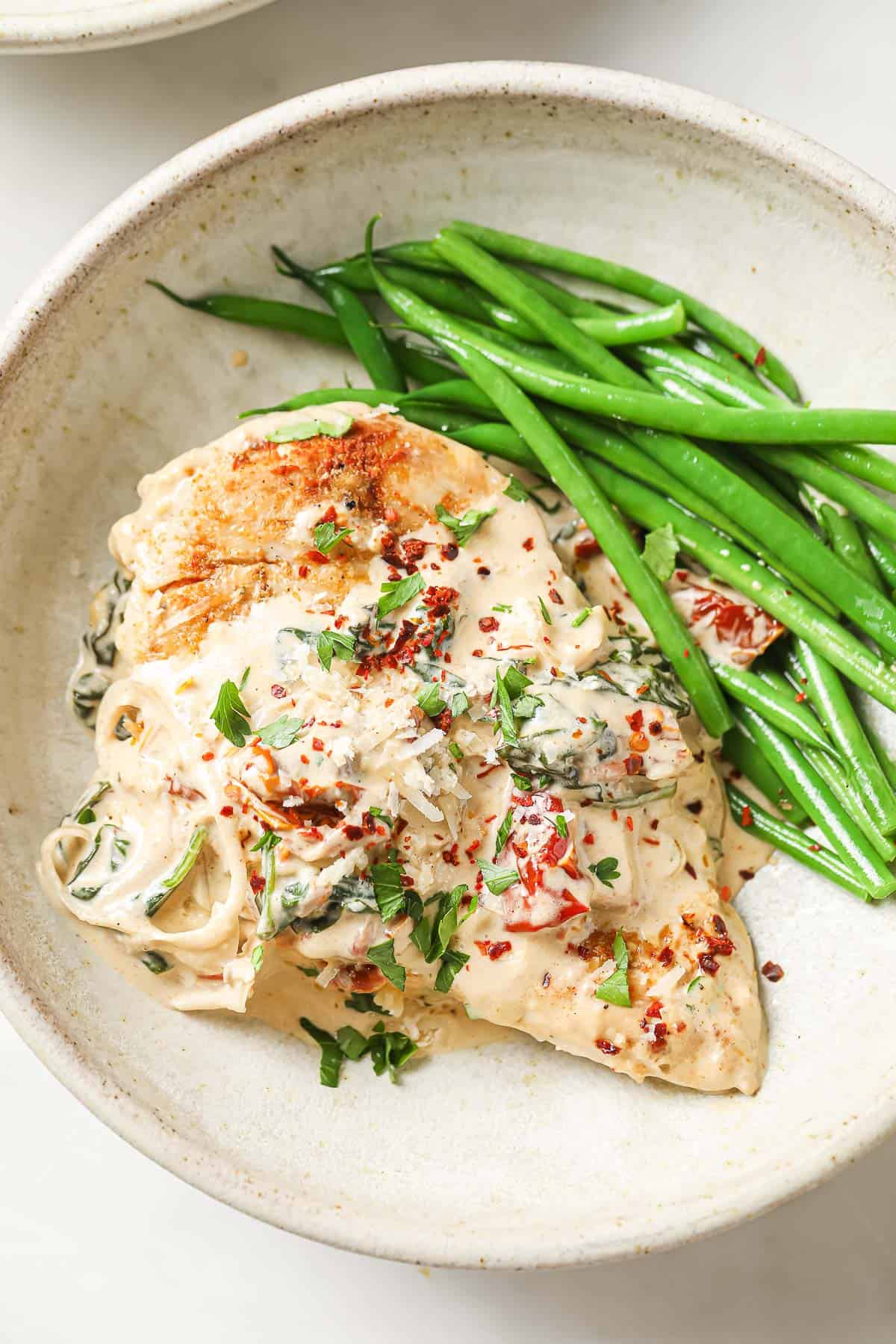 A white ceramic bowl with a chicken breast, topped with a sun-dried tomato cream sauce, with green beans on the side.