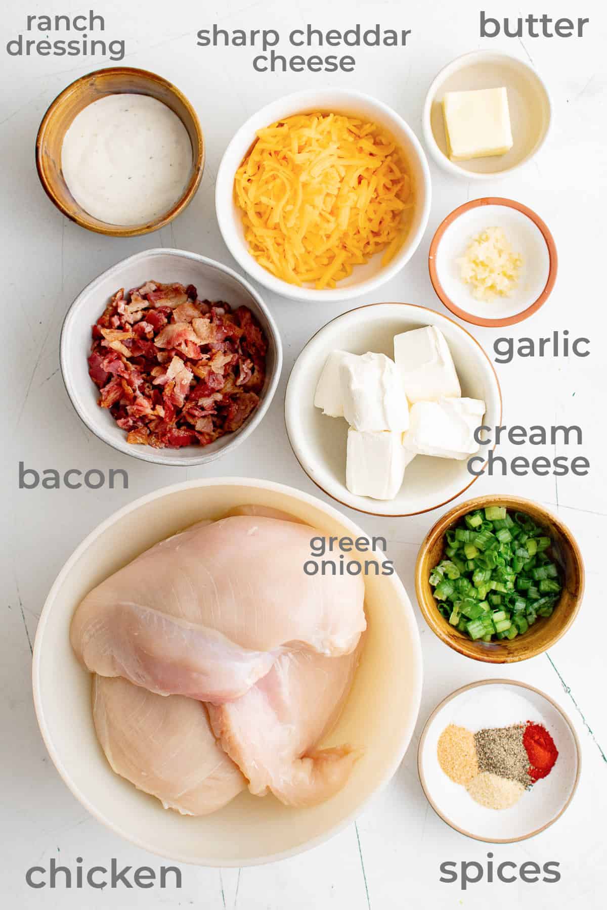 Ingredients laid out in individual bowls to make a stuffed chicken recipe - chicken, bacon, cream cheese, cheddar, ranch dressing, garlic, spices.