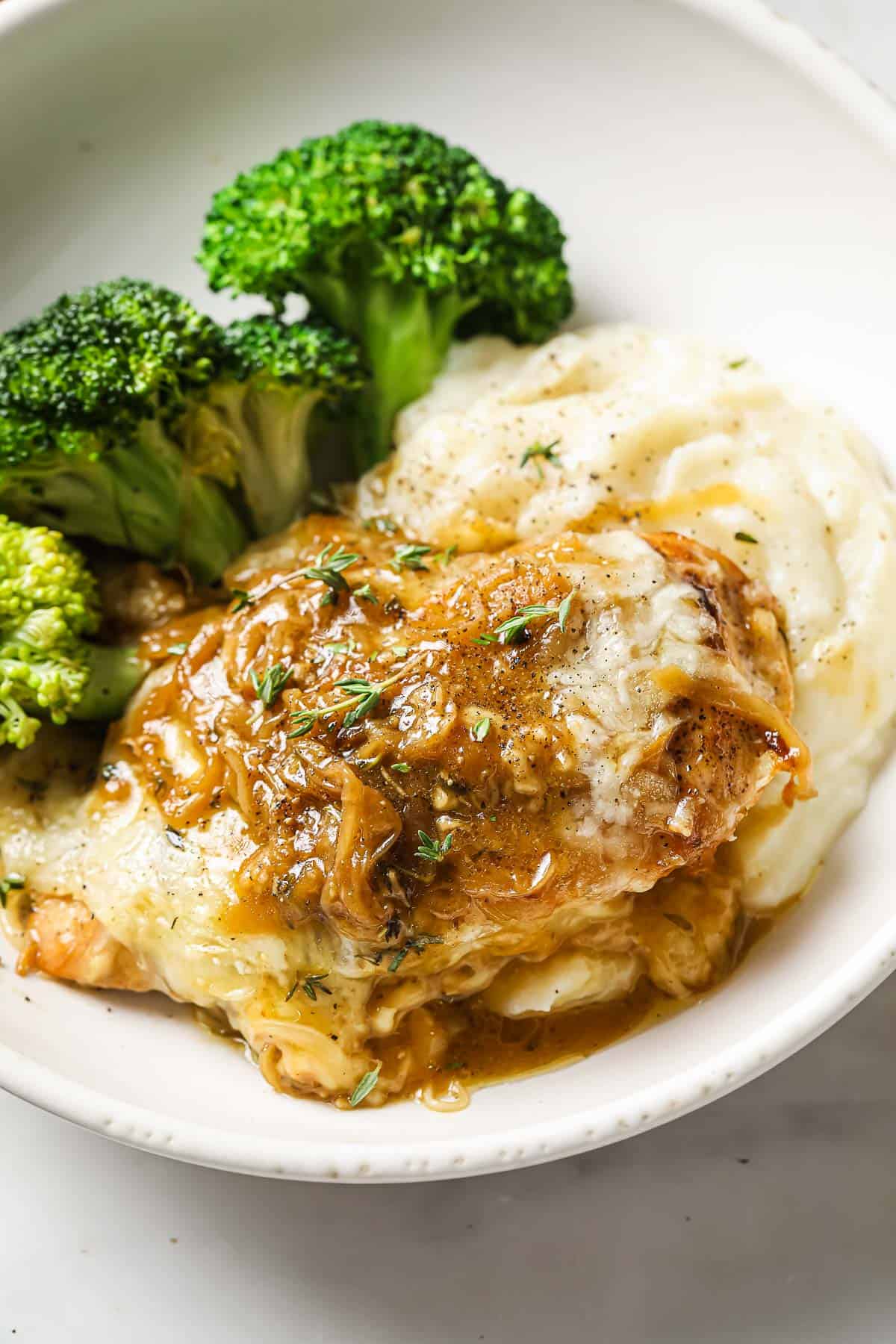 A white bowl with French onion chicken on top of cauliflower mash, served with steamed broccoli.
