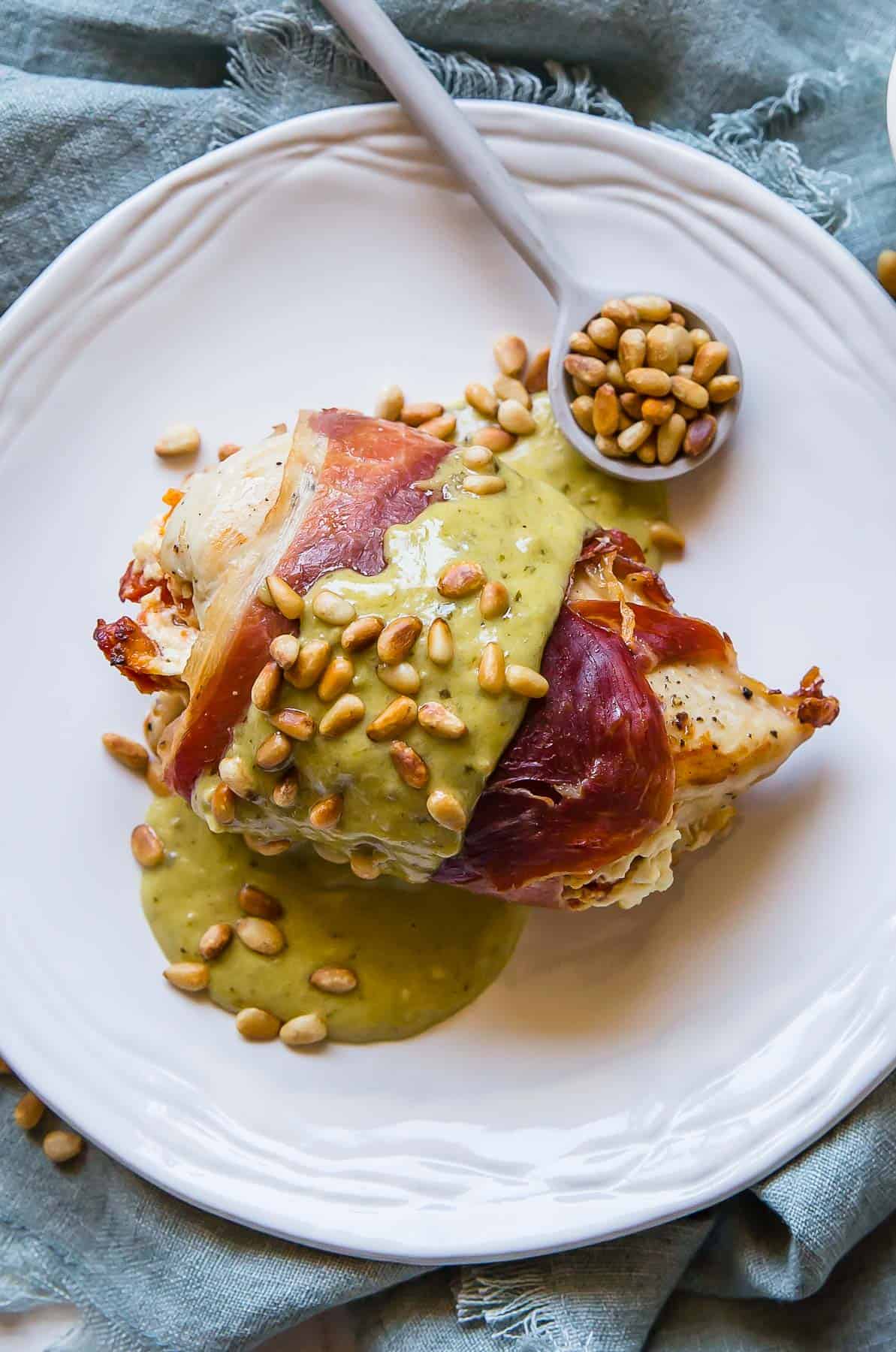 A serving plate with a chicken breast that is stuffed and then wrapped in prosciutto and topped with pesto cream sauce and toasted pine nuts.