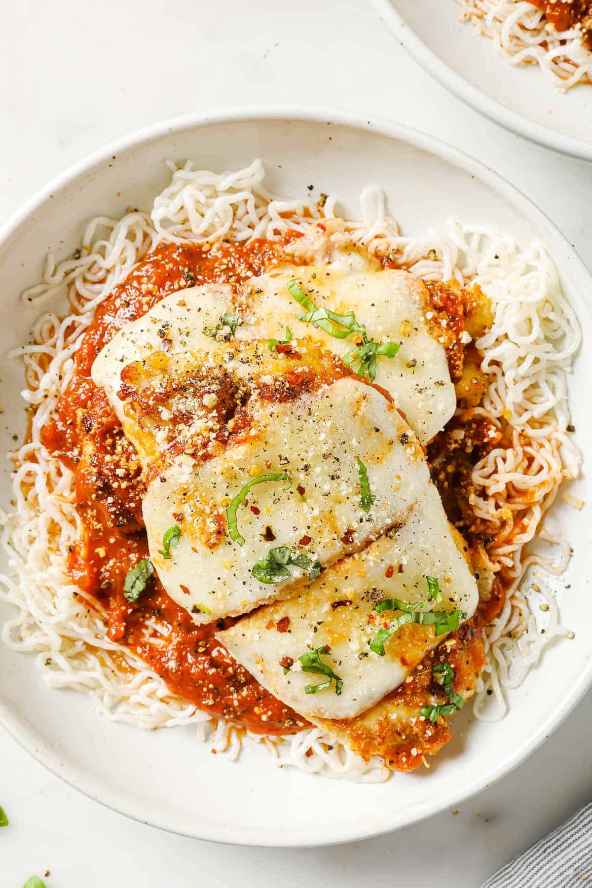 A white bowl with a plate of breaded chicken with melted cheese in a pool of marinara sauce, on top of shirataki noodles.