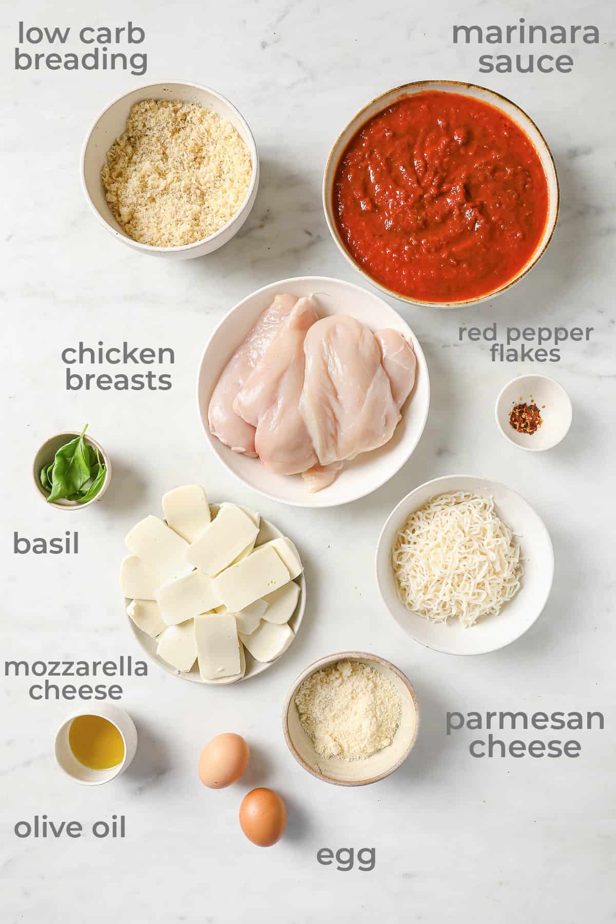 Ingredients laid out to make a low carb chicken parmigiana - chicken, marinara, cheese, basil, breading, egg, and olive oil.