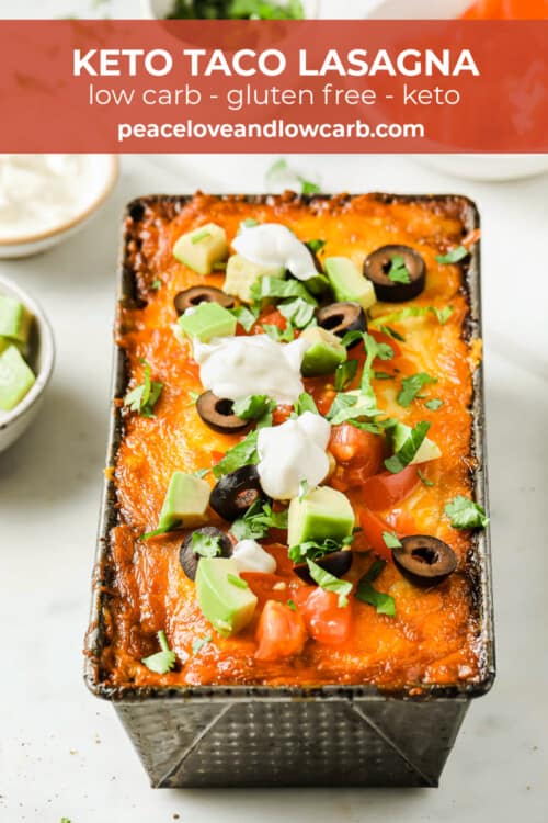 A loaf pan full of low carb lasagna, topped with golden brown baked cheese, olives, avocado, tomato, and sour cream.