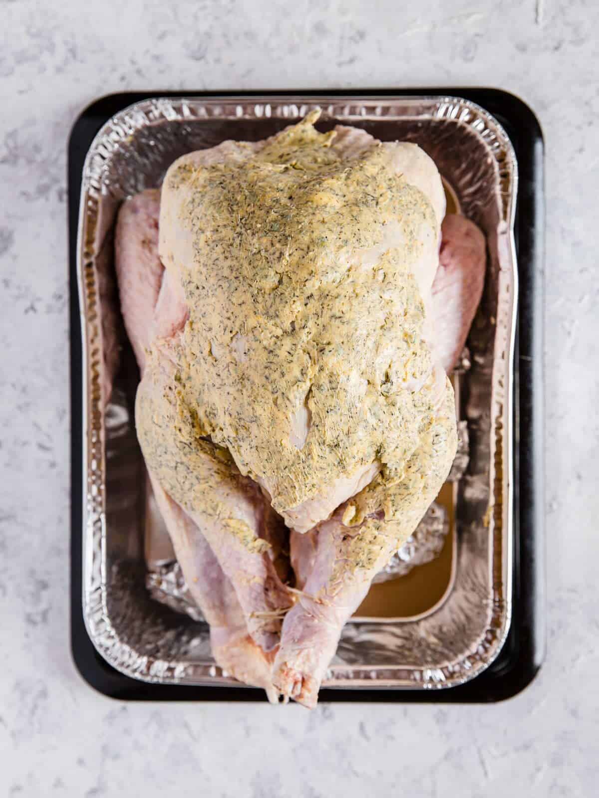 An aluminum roasting pan with a raw turkey in it that is covered with compound butter.