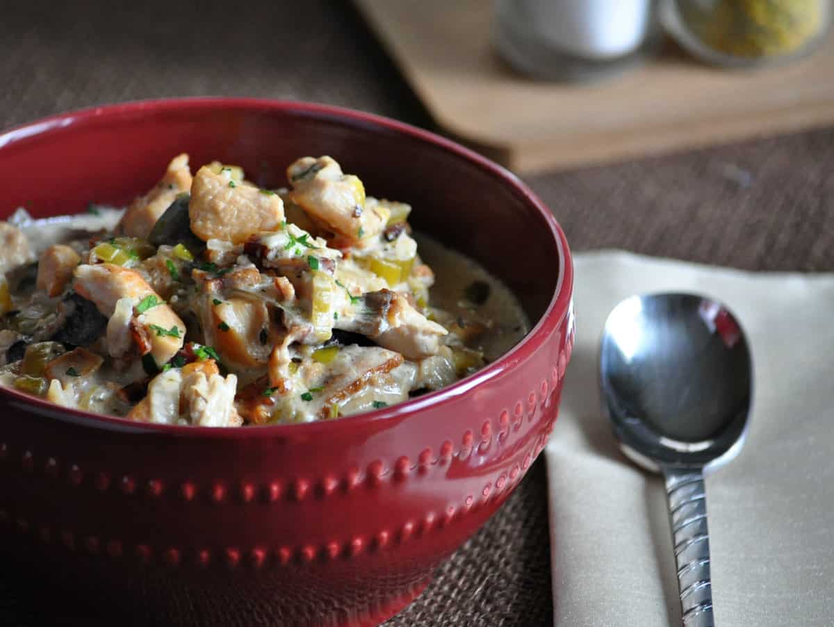 A red ceramic bowl full of a thick, creamy soup made with chicken, bacon and vegetables in a creamy broth.