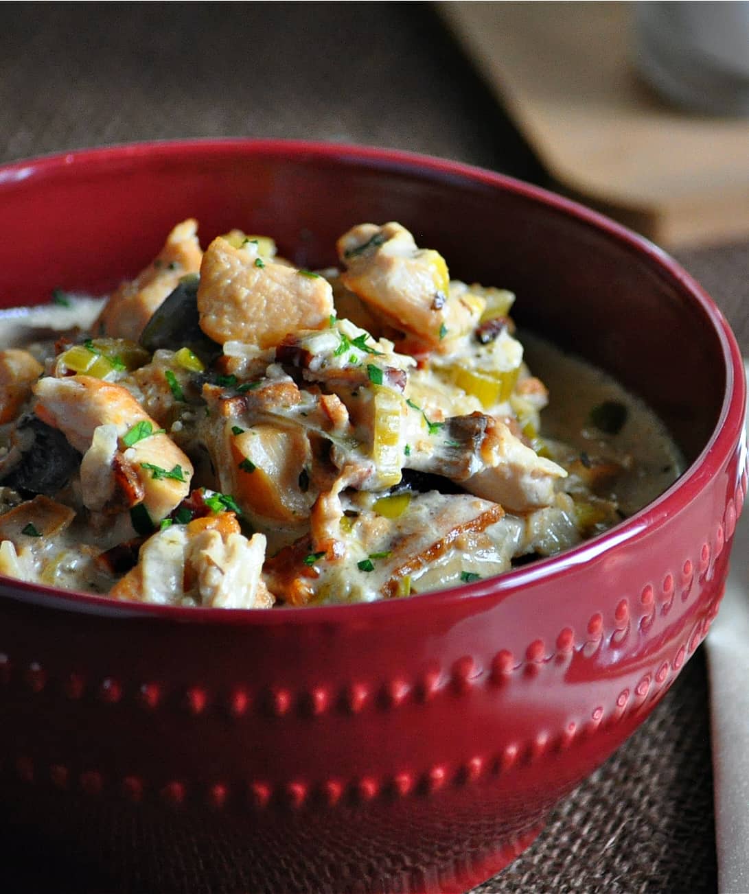 A red ceramic bowl full of a thick, creamy soup made with chicken, bacon and vegetables in a creamy broth.